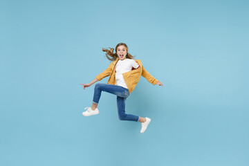 Full length portrait of excited little blonde kid girl 12-13 years old in yellow jacket isolated on blue background studio. Childhood lifestyle concept. Mock up copy space. Jumping, spreading hands.