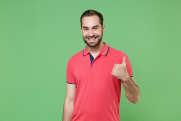 Smiling young bearded man guy in casual red pink t-shirt posing isolated on green wall background studio portrait. People sincere emotions lifestyle concept. Mock up copy space. Showing thumb up.