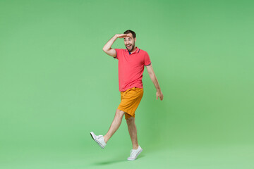 Fototapeta na wymiar Excited young bearded man guy in casual red pink t-shirt posing isolated on green background studio. People lifestyle concept. Mock up copy space. Holding hand at forehead looking far away distance.