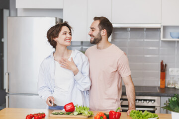 Laughing young couple friends guy girl in casual clothes preparing vegetable salad cooking food in kitchen at home. Dieting family healthy lifestyle concept. Mock up copy space. Looking at each other.