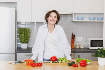 Smiling cheerful young brunette housewife woman girl in white casual clothes preparing vegetable salad cooking food in light kitchen at home. Dieting healthy lifestyle concept. Mock up copy space.