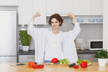 Smiling young housewife woman girl in casual clothes preparing vegetable salad cooking food in light kitchen at home. Dieting healthy lifestyle concept. Mock up copy space. Showing biceps, muscles.
