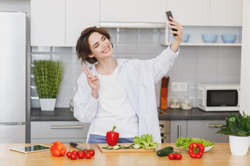 Smiling housewife woman in casual clothes preparing vegetable salad cooking food in light kitchen at home. Dieting healthy lifestyle concept. Doing selfie shot on mobile phone, showing victory sign.