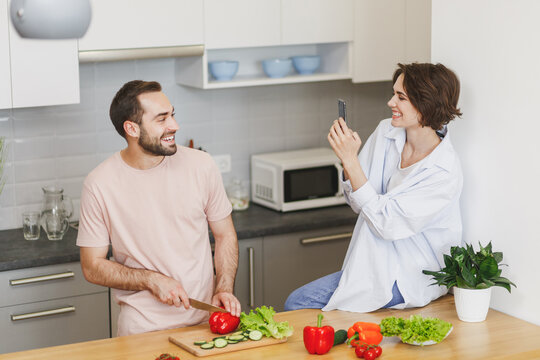 Funny couple friends guy girl sit on table preparing vegetable salad cooking food in light kitchen at home. Dieting family healthy lifestyle concept. Mock up copy space. Take pictures on mobile phone.