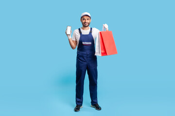Full length positive professional courier in uniform holding shopping bags and mobile phone with mock up blank display for advertise image, online delivery order at fashion store. studio shot isolated