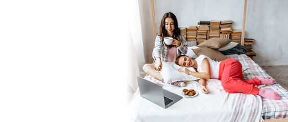 Two brunette girls are lying on the bed at home on the weekend with a laptop. Girlfriends have...