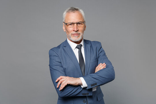 Confident elderly gray-haired mustache bearded business man in classic blue suit shirt tie posing isolated on grey background studio. Achievement career wealth business concept. Holding hands crossed.