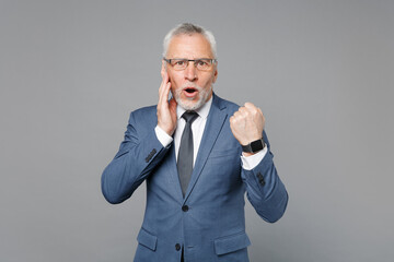 Shocked elderly gray-haired business man in classic blue suit shirt tie isolated on grey background studio. Achievement career wealth business concept. Wearing smart watch on hand, put hand on cheek.