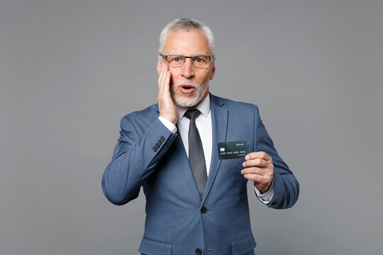 Amazed elderly gray-haired business man in classic blue suit shirt tie isolated on grey wall background studio. Achievement career wealth business concept. Hold credit bank card, put hand on cheek.