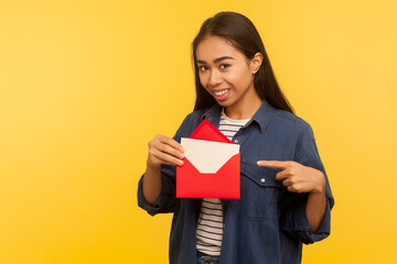 Fototapeta premium I got love letter on Valentine's day. Happy beautiful girl in denim shirt pointing letter in red envelope, holding greeting card and smiling joyfully. indoor studio shot isolated on yellow background