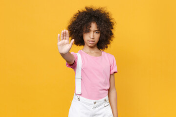 Little african american kid girl 12-13 years old in pink t-shirt isolated on yellow wall background studio portrait. Childhood lifestyle concept. Mock up copy space. Showing stop gesture with palm.