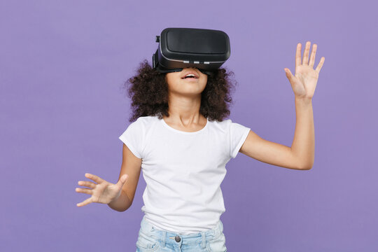 Shocked little african american kid girl 12-13 years old in white t-shirt isolated on violet background. Childhood concept. Watching in headset touch something push on button point at virtual screen. - Powered by Adobe