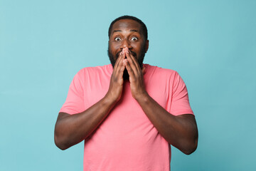 Amazed young african american man guy in casual pink t-shirt posing isolated on blue wall background studio portrait. People emotions lifestyle concept. Mock up copy space. Covering mouth with hands.