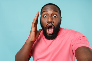 Close up of shocked young african american man guy in pink t-shirt isolated on blue wall background. People lifestyle concept. Mock up copy space. Doing selfie shot on mobile phone put hand on cheek.