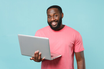 Smiling young african american man guy in casual pink t-shirt isolated on pastel blue wall background studio portrait. People lifestyle concept. Mock up copy space. Working on laptop pc computer.