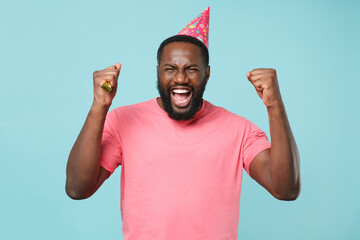 Joyful young african american man guy in casual pink t-shirt birthday hat isolated on pastel blue background studio portrait. People lifestyle concept. Mock up copy space. Hold pipe, clenching fists.