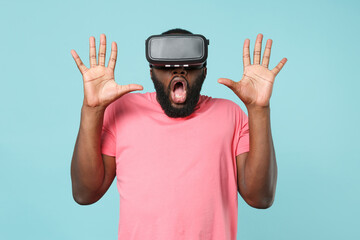 Shocked young african american man guy in casual pink t-shirt isolated on pastel blue background. People lifestyle concept. Mock up copy space. Watching in headset, rising hands up, showing palms.