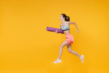Full length portrait beautiful young fitness woman in sportswear working out isolated on yellow wall background. Workout sport motivation lifestyle concept. Mock up copy space. Jumping, hold yoga mat.