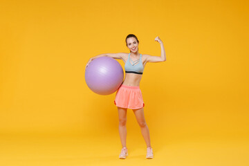 Full length portrait of smiling young fitness woman girl in sportswear working out isolated on yellow wall background. Workout sport motivation lifestyle concept. Hold fitball showing biceps, muscles.