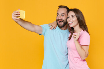 Cheerful couple two friends guy girl in blue pink t-shirts isolated on yellow background. People lifestyle concept. Mock up copy space. Doing selfie shot on mobile phone hugging showing victory sign.