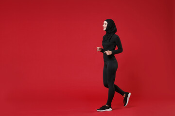 Full length portrait side view of smiling young arabian muslim woman in hijab black clothes posing isolated on red background. People religious lifestyle concept. Mock up copy space. Looking aside.