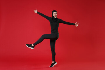 Full length portrait of excited young arabian muslim woman in hijab black clothes isolated on red background. People religious lifestyle concept. Mock up copy space. Rising speading hands and legs.