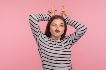 Portrait of carefree happy woman in striped sweatshirt winking playfully and showing bunny ears, aping with comical childish grimace, humorous expression. studio shot isolated on pink background