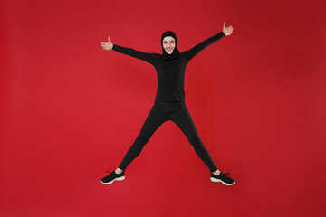 Fototapeta na wymiar Full length portrait of funny young arabian muslim woman in hijab black clothes isolated on red background. People religious lifestyle concept. Mock up copy space. Jumping, spreading hands and legs.