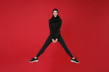 Fototapeta na wymiar Full length portrait of smiling young arabian muslim woman in hijab black clothes posing isolated on red background. People religious lifestyle concept. Mock up copy space. Jumping, spreading legs.