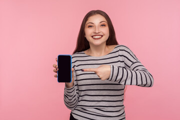 Look at device advertisement! Portrait of happy smiling woman in striped sweatshirt pointing mobile phone with empty display, mock up for app promotion, online web service. indoor studio shot isolated