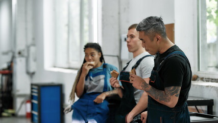 Obraz premium Lunch time. Team of diverse mechanics in uniform, two men and a woman eating sandwiches, while having a break at auto repair shop