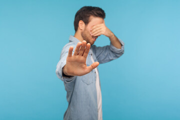 No, don't want to watch! Portrait of confused shy man in worker denim shirt covering eyes showing...