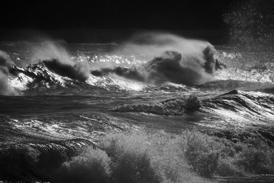 A stormy sea with gale force winds and the high tide creates huge waves which crash on the rocky beach. The setting sun glints across the water and the spray creates a mist across the water
