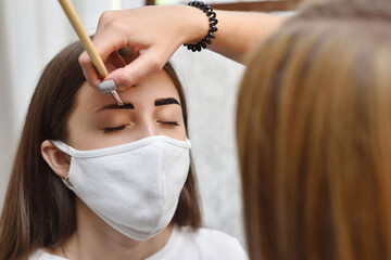 A young girl in a protective mask does henna eyebrow coloring in a beauty salon during the covid-19...