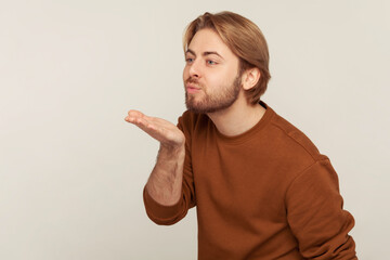 Love you, darling! Portrait of handsome man with neat hair and beard wearing sweatshirt, sending romantic air kiss, flirting expressing fondness and affection. studio shot isolated on gray background