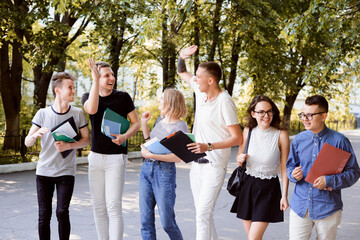 Group of laughing students with books, folders and other studying stuff outdoors on a bright sunny day; park and building of university on background. All friends wear casual clothes, girls carry bags