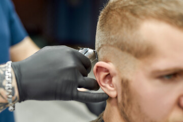 Visiting barbershop. Close up shot of a young caucasian man getting trendy haircut. Barber using professional electric hair clipper, serving client