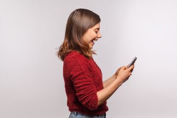 Side view, amazed brunette girl in shaggy sweater using mobile phone with surprised expression, typing or reading shocking message on social network, online communication. studio shot isolated on gray