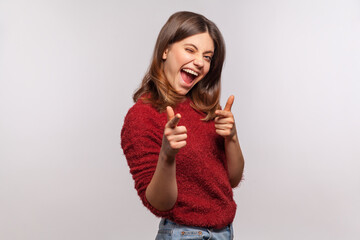 Hey you! Portrait of playful brunette woman in shaggy sweater pointing finger to camera, smiling excitedly, winking and flirting, choosing lucky winner. indoor studio shot isolated on gray background