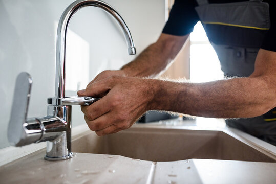 Close up of hands of aged repairman in uniform working, fixing broken kitchen tap using adjustable wrench. Repair service concept