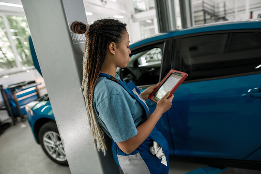 Young african american woman, professional female mechanic using tablet pc, while standing near blue car at auto repair shop