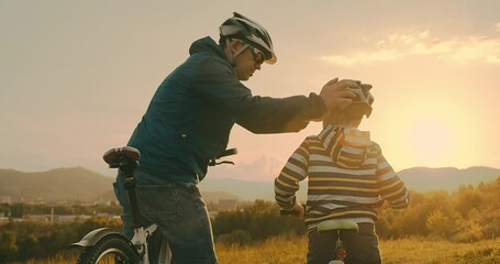 Father and son ride a bike. Happy cute boy in helmet learn to riding a bike in park on green meadow in summer day at sunset time. Family weekend. 4K video, Slow motion
