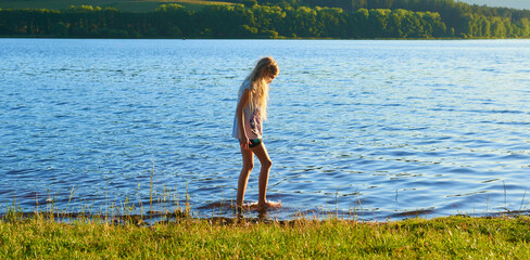 Alone child blond girl standing in the water in the light of the setting sun in summer