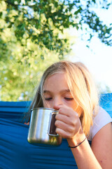 Child girl lying in a hammock and drinking water from a tin cup