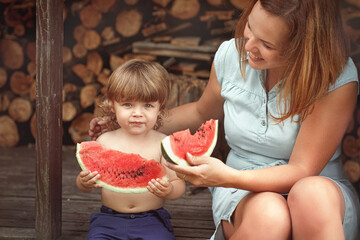 happy family little boy 2 years holds in hands juicy red watermelon on yard sitting near woodpile of firewood in village with mother looks at him