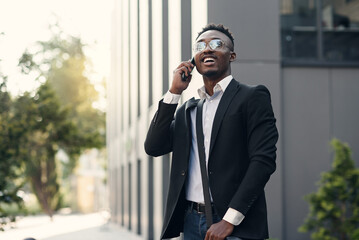 Serious young businessman in black suit standing and talking on cell phone near business center