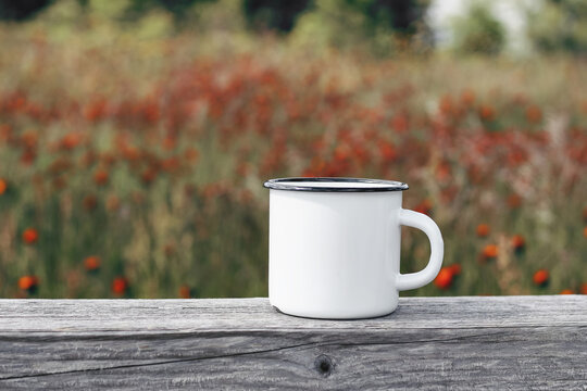 Close up of metal mug on old wooden table, board with defocused blooming mountain meadow. Outdoor tea, coffee time. Mockup of white enamel cup. Lifestyle relax, trekking and camping concept.