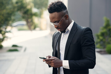 Serious black businessman texting on smartphone outdoors. Young african-american salesman working with mobile near business centre on the bench in urban area cityscape