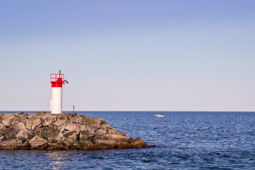 Red and white lighthouse on a rocky pier amid blue stretch of water with a white boat and clear blue sky in bright sunny summer day. Blue sky and calm water, horizontal orientation
