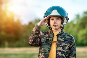 A boy in a pilot's helmet on a background of greenery salutes. Dream concept, choice of profession, military service. Copy space.
